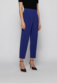 Pantalons ajustés bleu vif avec plis, taille haute et jambes fuselées, associés à des talons aiguilles noirs avec lanières autour de la cheville.