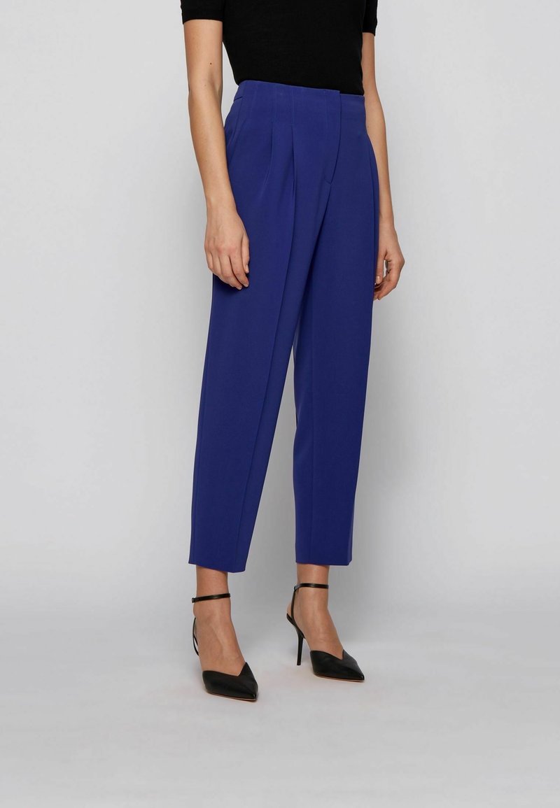 Pantalons ajustés bleu vif avec plis, taille haute et jambes fuselées, associés à des talons aiguilles noirs avec lanières autour de la cheville.