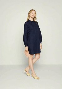 Mulher com um vestido camiseiro de mangas longas azul-marinho, segurando um acessório de pele rosa, usando sandálias de salto amarelas, de pé contra um fundo simples.