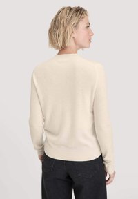 Cremfarbener Pullover aus weichem Stoff, mit einem runden Ausschnitt, gerippten Bündchen und Saum, getragen mit dunklen Jeans, von hinten gesehen.