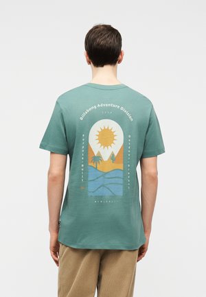 Mees, kes kannab türkiissinist T-särki, mille seljal on kõrbemaastiku pilt päikese, kaktuse, palmi ja mägedega.