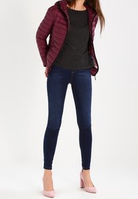 Burgundowa pikowana kurtka z kapturem, zestawiona z czarną koszulą, ciemnoniebieskimi wąskimi dżinsami i jasnoróżowymi szpilkami. Gładkie tekstury na całej powierzchni.