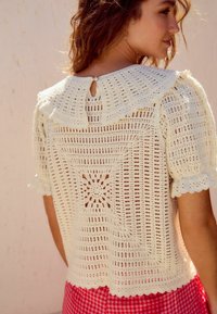Haut au crochet crème avec un col festonné, des manches courtes bouffantes, et des détails en dentelle complexes, présentant une fermeture en fente au dos.