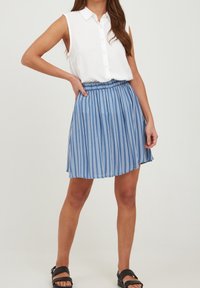 Blå och vit randig kjol med en plisserad design, kombinerad med en ärmlös vit skjorta med knäppning och svarta sandaler.