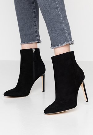 Bottes chevilles en daim noir à bout pointu avec fermetures éclair latérales et talons aiguilles hauts portées avec un jean gris à ourlet effiloché.