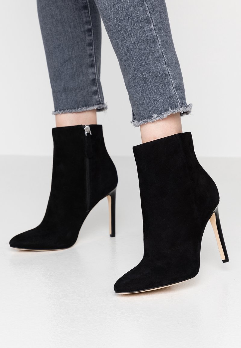 Bottes chevilles en daim noir à bout pointu avec fermetures éclair latérales et talons aiguilles hauts portées avec un jean gris à ourlet effiloché.