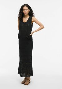 Robe en tricot noir au design de débardeur, présentant une texture de tricot ouvert et une silhouette ajustée. Accentuée par une ceinture.