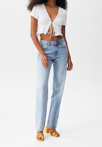 Top blanc coupé avec un nœud à l'avant, jean droit bleu clair, et sandales à talons blocs jaunes avec des détails de lanières.