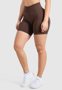 Bruine sportieve shorts met een hoge tailleband en een aansluitend ontwerp. Glad textuur met subtiele zijpaneel details. Draagt met witte sneakers.