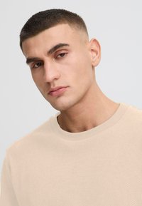 Beige strukturierter Pullover mit Rundhalsausschnitt. Der Stoff hat ein leichtes Muster. Das Model hat kurze Haare und einen neutralen Gesichtsausdruck.