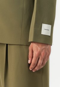 Olive green blazer sleeve features a smooth texture and a white label with "Calvin Klein" in black text. A well-fitted design accentuates the arm.