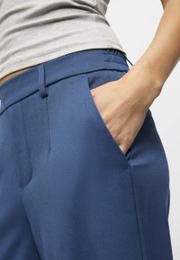 Námořnické modré kalhoty na míru s bočními kapsami, vyrobené z hladké, texturované látky a s přilnavým pasem. Viditelná ruka v kapse.