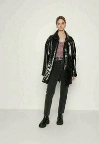 Mulher em pé vestindo uma jaqueta de couro preta brilhante, blusa padrão em cor maroon, calças pretas de cintura alta, cinto preto e botas de tornozelo pretas, em frente a uma parede lisa.