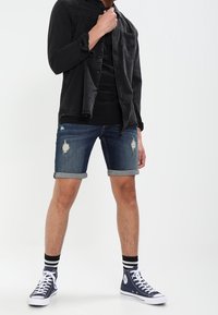 Jeune homme portant une chemise noire par-dessus un t-shirt noir, un short en denim déchiré et des baskets avec des chaussettes rayées, posé devant un fond neutre.