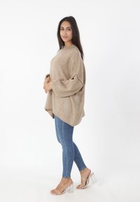 Beige oversized gebreide trui met ballonmouwen, gecombineerd met blauwe skinny jeans en transparante sandalen met blokhak. Eenvoudig en textuurrijk ontwerp.