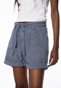 Un short en coton gris présente une taille élastique avec cordon de serrage, des poches latérales et des poignets retroussés. Le matériau a une texture douce.