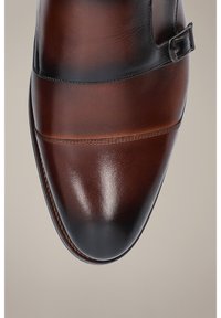 Brązowy skórzany but z polerowanym wykończeniem, z eleganckim wzornictwem i czarnym akcentem na środku, zabezpieczony klamrą.