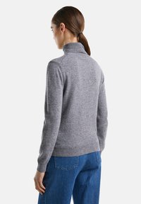 Donna con i capelli castani legati in una coda di cavallo, che indossa un maglione grigio a collo alto e jeans blu, vista da dietro su uno sfondo bianco.