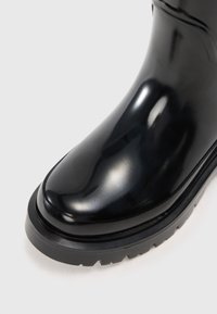 Bottine en caoutchouc noire avec une finition brillante, bout rond, semelle noire texturée et détail de couture subtil le long du bord supérieur.