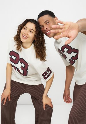 Deux jeunes adultes portant des chemises blanches assorties avec le numéro 23 et des pantalons marron à fines rayures, souriants et se penchant ensemble vers l'avant.