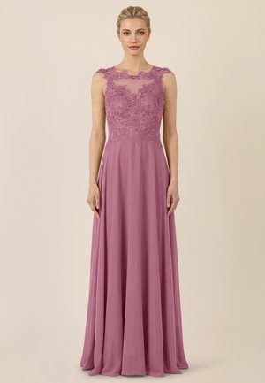 Woman wearing a floor-length mauve gown with floral lace bodice and sheer neckline, standing against a plain beige background.