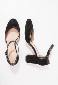 Černé vysoké podpatky z semišového materiálu, s designem peep-toe, s páskem kolem kotníku a texturovanými černými lakovými detaily na podpatku.