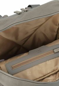 Sac à dos en tissu gris avec un intérieur en textile beige, comprenant un compartiment zippé et une doublure embossée du logo. Surface lisse et design structuré.