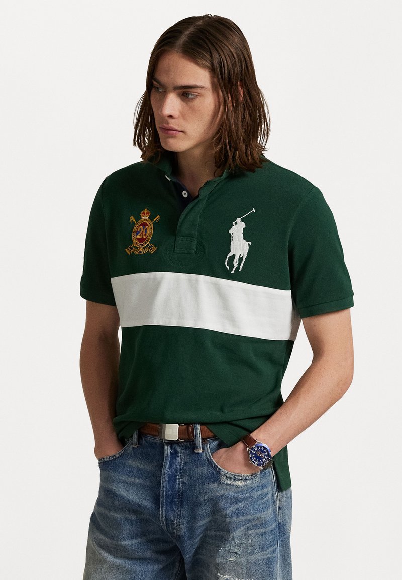 Zaļš polo krekls ar baltu horizontālu svītru, ar izšūtu ģerboni un balto polo spēlētāja logo. Saskaņots ar nolietotām zilām džinsām.