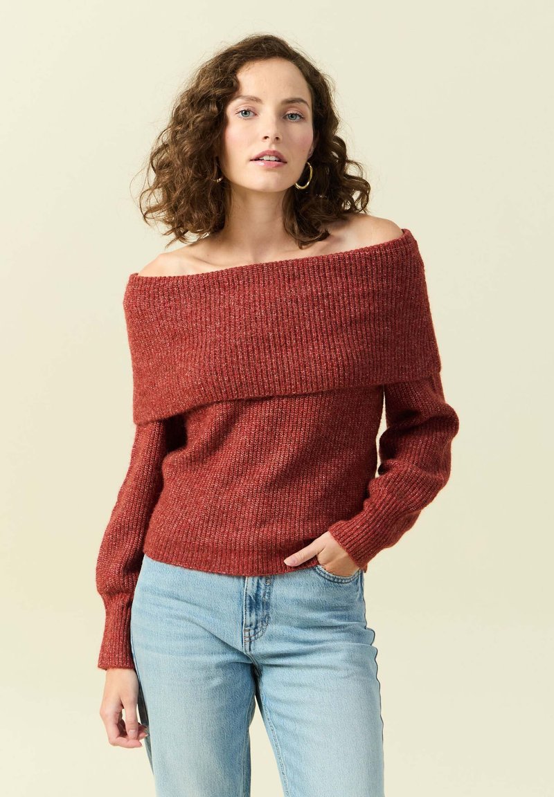 Maglione a spalle scoperte in ruggine con texture a costine e maniche lunghe, abbinato a jeans in denim azzurro chiaro.