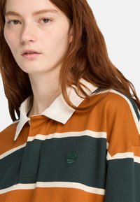 Młoda kobieta z długimi rudymi włosami, nosząca w paski pomarańczową, zieloną i białą koszulę z kołnierzykiem oraz małym logo drzewka na klatce piersiowej.