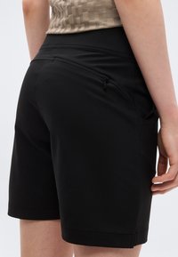 Κοντινό πλάνο ατόμου που φοράει μαύρο σορτς μέχρι το γόνατο με φερμουάρ στην πίσω τσέπη και μπεζ τοπ με σχέδια, σε λευκό φόντο.