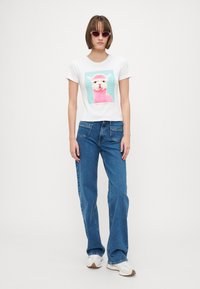 T-shirt blanc à manches courtes avec un graphisme coloré d'alpaga, associé à un jean flare bleu et des baskets blanches avec des semelles marron clair.