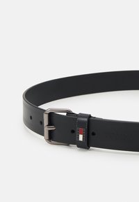 Cinturón de cuero negro con un acabado liso, hebilla de metal en tono plateado y una pequeña etiqueta de marca. Presenta múltiples agujeros para ajuste de tamaño.