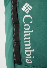 Sac à dos en tissu teal mettant en avant un logo blanc proéminent et des sangles noires avec des coutures blanches, présentant un design durable et texturé.