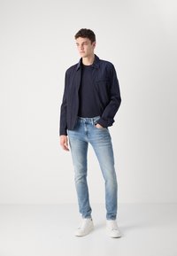 Chaqueta texturizada en azul marino con cuello, llevada sobre una camiseta oscura, combinada con jeans azul claro y zapatillas blancas. Diseño minimalista, corte moderno.