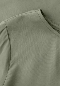 Nahaufnahme von olivgrünem Stoff, der die Naht und den Ausschnittsaum eines schlichten kurzärmeligen Kleidungsstücks zeigt.