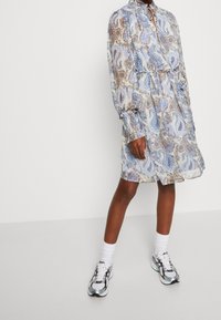 Robe paisley florale en bleu clair et crème, avec des manches longues et un col. Dotée d'une taille élastique et d'un tissu transparent.