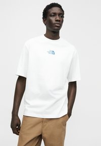 Λευκό βαμβακερό μπλουζάκι με κοντά μανίκια και στρογγυλή λαιμόκοψη. Διαθέτει μπλε λογότυπο στο στήθος, συνδυασμένο με καφέ υφασμάτινο παντελόνι με υφή.