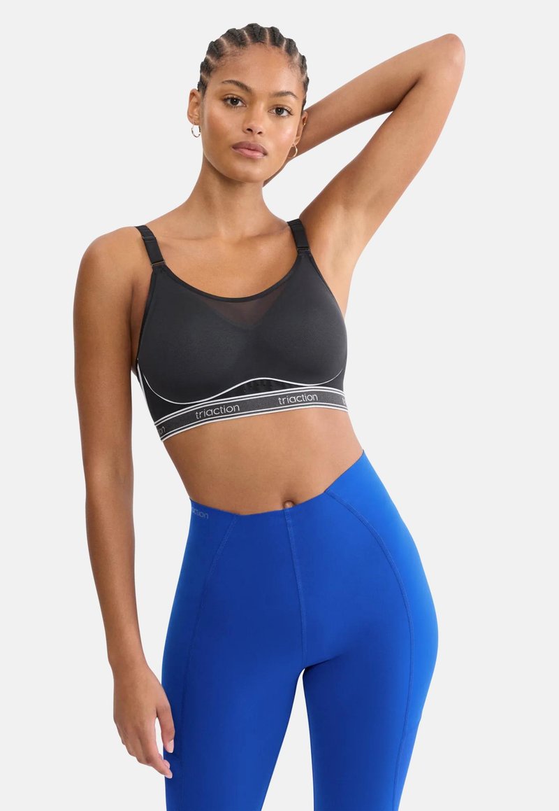 Γυναίκα με πλεγμένα μαλλιά που φοράει μαύρο αθλητικό σουτιέν και μπλε κολάν με ψηλή μέση, στέκεται με το ένα χέρι σηκωμένο και το άλλο χαλαρό.