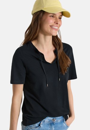 Smilende kvinde iført en gul kasket, sort kortærmet top med snore og lyseblå jeans, stående med den ene hånd i lommen.