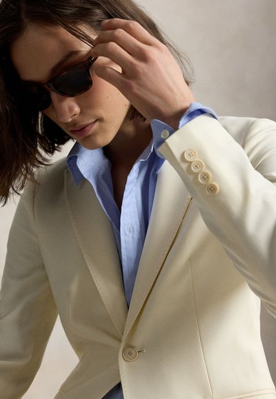 Vállig érő sötét hajú személy fekete napszemüveget, világoskék inget és krémszínű blézert visel, és jobb kezével igazítja a napszemüveget.