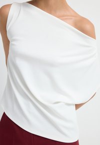 Top bianco off-shoulder, tessuto drappeggiato con una consistenza morbida, presenta un colletto diagonale. Abbinato a un pantalone bordeaux, mette in risalto curve sottili.