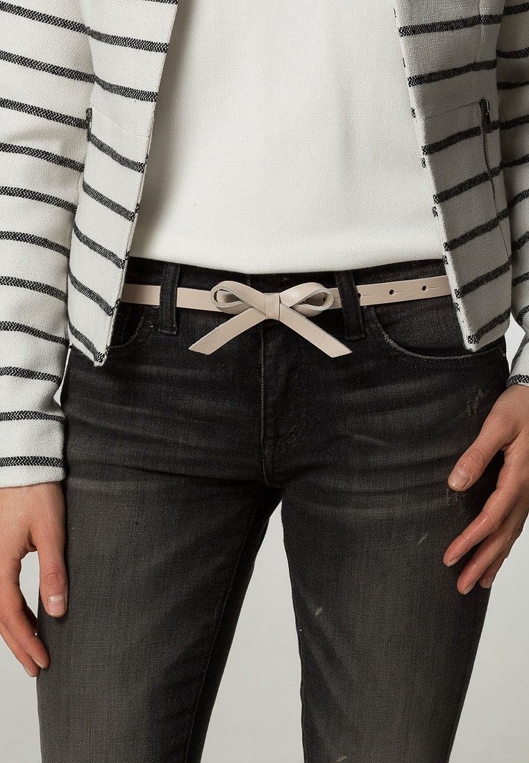 Personne portant un jean noir avec une ceinture nœud beige, un haut blanc et une veste blanche à rayures horizontales noires. Mains détendues sur les côtés.