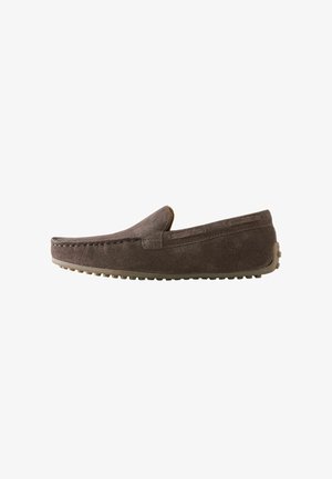 Mocasín marrón de ante sin cordones con suela de goma con tachuelas y detalles cosidos a lo largo de los bordes, mostrado de lado sobre un fondo blanco.