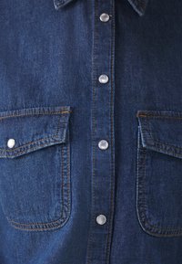 Camisa de mezclilla azul oscuro en la parte delantera con botones a presión de metal y dos bolsillos en el pecho con solapas y costuras naranjas en contraste.