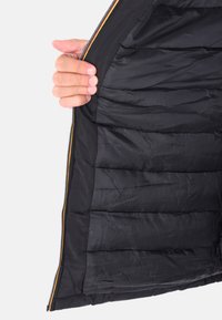 Una mano tiene aperto l'interno di una giacca nera trapuntata, mostrando la foderatura interna e il bordo della cerniera con bordi rossi e gialli.