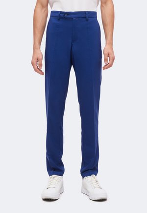 Homme portant un pantalon bleu droit et des baskets blanches, debout devant un fond uni.
