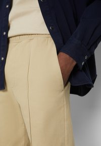 Chemise bleue à boutons superposée sur un pantalon beige à taille élastique et plis, avec une main dans la poche, mettant en valeur la texture du tissu et les détails des coutures.
