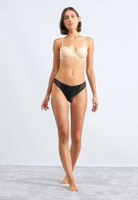 Beige gewatteerde beha met dunne straps, gecombineerd met een zwarte doorschijnende string. Glad fabric en minimalistisch ontwerp. Model gepresenteerd op een neutrale achtergrond.