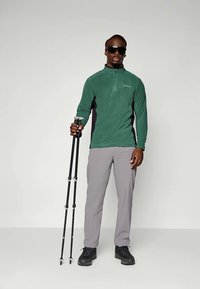 Green fleece zip-up top with black side panels, gray pants, and black hiking boots. Person holds trekking poles and wears sunglasses.